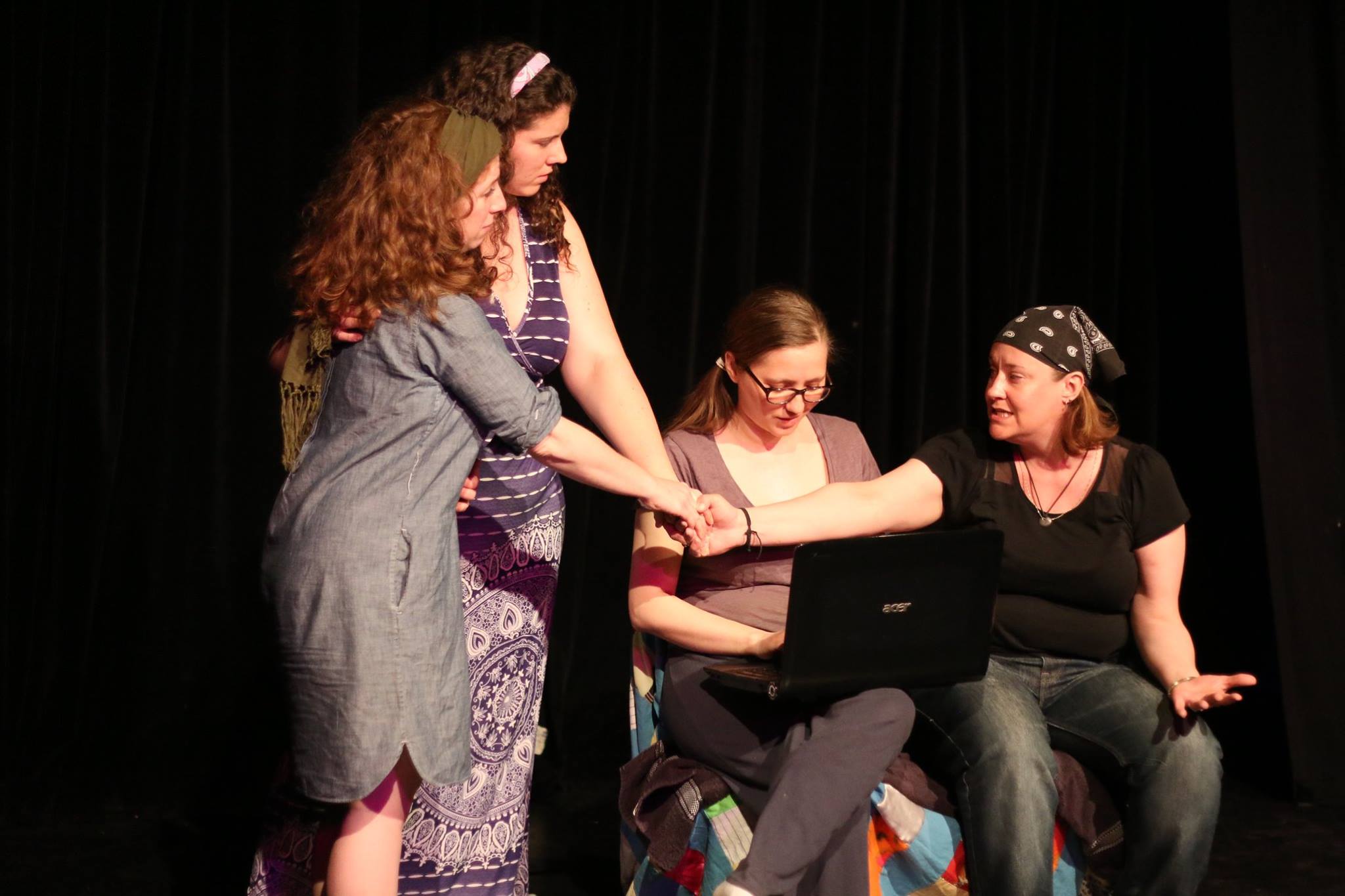 Three women characters in casual counterculture clothing, join hands while another character works on a laptop